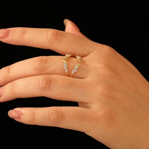 Pu&ograve; includere: Un anello dorato dal design unico, con piccole pietre preziose scintillanti. L'anello ha un gambo diviso e una parte superiore decorativa con un gruppo di pietre. L'anello &egrave; mostrato su una mano su sfondo nero.