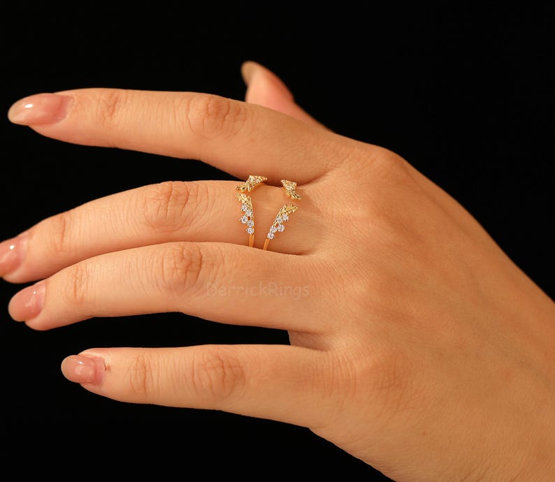 Pu&ograve; includere: Un anello dorato dal design unico, con piccole pietre preziose scintillanti. L'anello ha un gambo diviso e una parte superiore decorativa con un gruppo di pietre. L'anello &egrave; mostrato su una mano su sfondo nero.