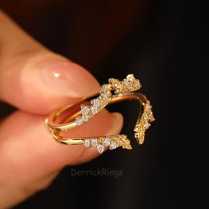 Pu&ograve; includere: Un anello color oro con un design a foglie e piccole gemme trasparenti. L'anello ha una doppia fascia e un design diviso. Il testo "DerrickRings" &egrave; visibile.