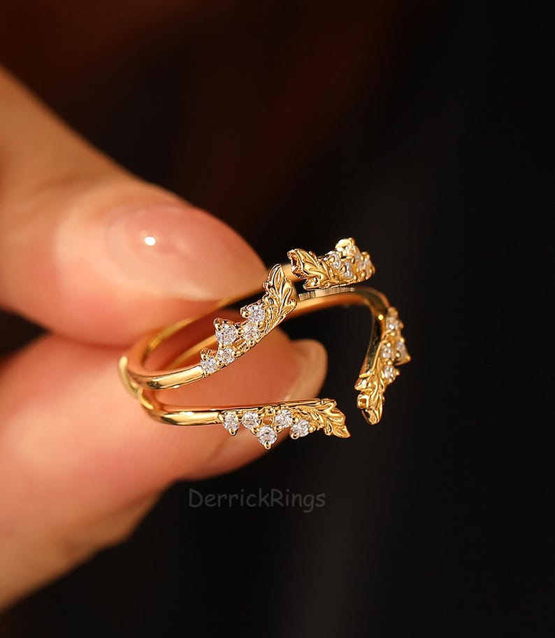 Pu&ograve; includere: Un anello color oro con un design a foglie e piccole gemme trasparenti. L'anello ha una doppia fascia e un design diviso. Il testo "DerrickRings" &egrave; visibile.