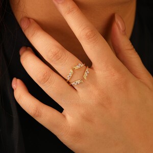 Pu&ograve; includere: Un anello color oro dal design unico. L'anello presenta una doppia fascia con una forma geometrica al centro, ornata da piccole pietre scintillanti. L'anello &egrave; progettato per essere indossato attorno a un anello esistente.