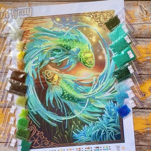 Op de afbeelding: Een close-up van een diamant schilderset met een ontwerp van twee blauwe en groene vissen die in een cirkel zwemmen. De set bevat een canvas, kralen en instructies. De kralen zijn georganiseerd in kleine plastic zakjes die zijn gelabeld met nummers.