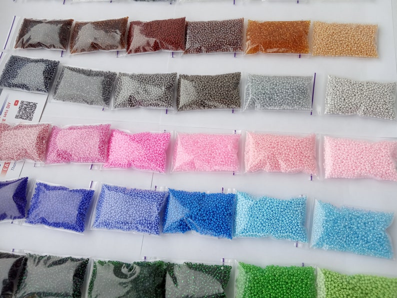 May include: Assortment of small, colorful beads in clear plastic bags. The beads are in various shades, including brown, gray, pink, blue, and green. The bags are arranged in neat rows, suggesting a craft supply or DIY project.
