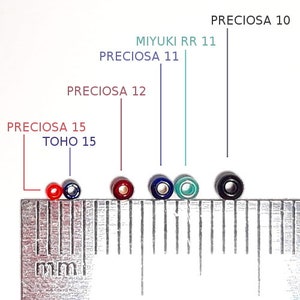 May include: A collection of colorful seed beads, including red, blue, green, and black, are displayed next to a metric ruler. The beads are labeled with brand names and sizes, such as Preciosa and Miyuki.