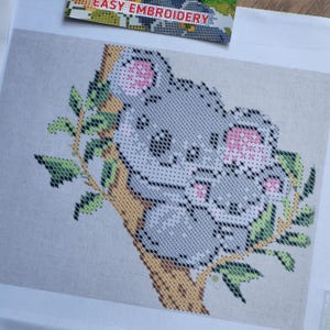 以下が含まれることがあります： 緑の葉が付いた木の枝にいるコアラと赤ちゃんコアラのクロスステッチ刺繍。コアラはグレーで、耳と鼻はピンク色です。「EASY EMBROIDERY」の文字が上部に表示されています。