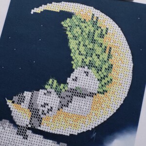 May include: Cross-stitch art featuring a crescent moon with a yellow and green design. Two panda bears are nestled on the moon, with a dark blue background and small white stars. The artwork is a close-up shot.