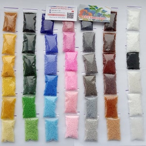 May include: A collection of small, colorful beads in clear plastic bags. The beads are in a variety of colors, including red, yellow, green, blue, pink, and purple. The bags are arranged in rows and columns, creating a visually appealing display. The image also includes a small card with text.