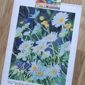 以下が含まれることがあります： 白いヒナギク、黄色の中心、緑の葉のフローラルデザインのクロスステッチ刺繍キット。青い蝶と黄色の太陽も含まれています。キットには「EASY EMBROIDERY」と表示されています。