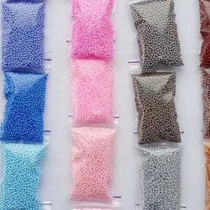 May include: Rows of small, colorful beads in clear plastic bags. Colors include red, yellow, green, blue, pink, gray, brown, black, white, and purple. The beads appear to be uniform in size and shape, suitable for crafting or jewelry making.