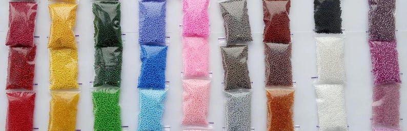 May include: Rows of small, colorful beads in clear plastic bags. Colors include red, yellow, green, blue, pink, gray, brown, black, white, and purple. The beads appear to be uniform in size and shape, suitable for crafting or jewelry making.