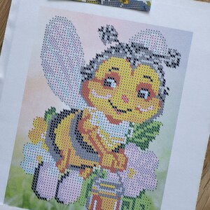 以下が含まれることがあります： 黄色、黒、白の縞模様の、笑顔の漫画のミツバチのクロスステッチパターン。ミツバチはピンクの花と緑の葉に囲まれ、蜂蜜の壺があります。背景はピンクと緑の柔らかなグラデーションです。