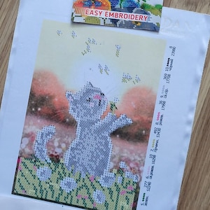 以下が含まれることがあります： 花畑にいる灰色の猫を描いたクロスステッチキット。猫はピンクの鼻をしており、小さな黄色の花に向かって手を伸ばしています。背景は柔らかいオレンジと黄色のグラデーションです。