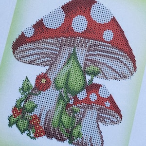 Op de afbeelding: Kruissteekontwerp met twee rode paddenstoelen met witte stippen, groene bladeren en kleine rode bloemen. De paddenstoelen staan centraal, met gedetailleerde steken die een textuur creëren. Het ontwerp staat op een witte achtergrond.