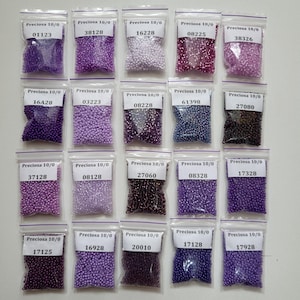 May include: An assortment of small glass beads in clear plastic bags. Each bag is labelled with "Preciosa 10/0" and a unique number. The beads are various shades of purple, from pale lilac to dark violet.