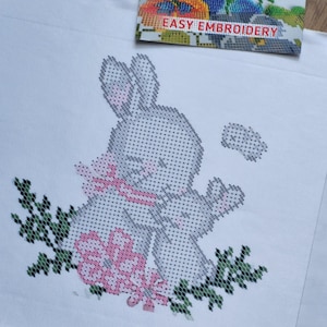 以下が含まれることがあります： ピンクのリボンとピンクの花を持つ灰色のウサギのクロスステッチパターン。緑の葉で囲まれています。「EASY EMBROIDERY」の文字が画像上部にあります。