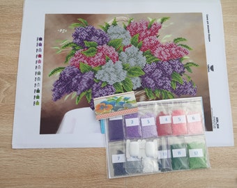 May include: A close-up of a diamond painting kit featuring a picture of a bouquet of purple, pink, and white lilacs in a wicker basket. The kit includes 12 bags of colourful beads, a small instruction sheet, and a white canvas with a printed design.
