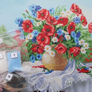 Può includere: Un disegno floreale a punto croce con un bouquet di papaveri rossi, fiordalisi blu e margherite bianche in un vaso. L'immagine include piccoli sacchetti di perline di vari colori e una bobina di filo.