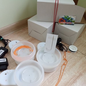 May include: A set of white plastic electric bead spinners with power adapters, orange beads, a needle, and thread. The equipment is packaged in white cardboard boxes, one featuring a small card with a floral image and the text EASY EMBROIDERY.