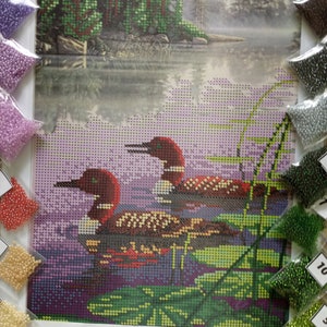 May include: A diamond painting kit featuring two red-headed loons swimming in a lake with green lily pads and a purple and green background. The kit includes a variety of colored beads in small plastic bags.