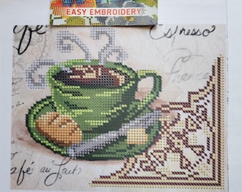 Op de afbeelding: Kruissteekpatroon met een groene koffiekop met stoom, een schotel en een koekje. De achtergrond heeft handschrift en een decoratieve hoek. De tekst "Easy Embroidery" staat bovenaan.