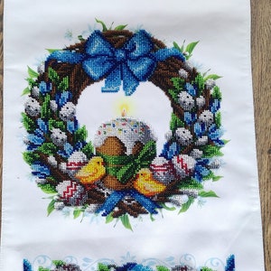 May include: A white fabric Easter wreath with a blue bow, decorated with green leaves, white flowers, and colourful Easter eggs. A brown Easter cake with white icing and a lit candle is in the centre of the wreath. Two yellow chicks are on either side of the cake.