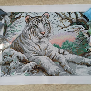 May include: A white tiger with black stripes is lying down in a snowy forest setting. The tiger is surrounded by trees and bushes. The background is a soft pink and blue sunset. The image is a diamond painting kit with numbered bags of beads.
