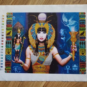 Op de afbeelding: Een kruissteekkunstwerk dat een Egyptische koningin afbeeldt met een uitgebreide gouden en blauwe hoofdtooi en sieraden. Het kunstwerk heeft een blauwe achtergrond met extra Egyptische figuren en symbolen en een rand met kleurstalen.