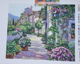 Puede incluir: Un proyecto de bordado parcialmente terminado que representa una escena de pueblo europeo con una calle adoquinada, edificios y flores vibrantes. El texto "EASY EMBROIDERY" es visible. El proyecto se asegura con un clip magnético.