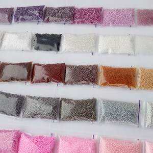 May include: Assortment of small, colorful beads in clear plastic bags. The beads are in various shades, including purple, white, black, brown, and gold. The bags are arranged in rows, creating a visually organized display of craft supplies.