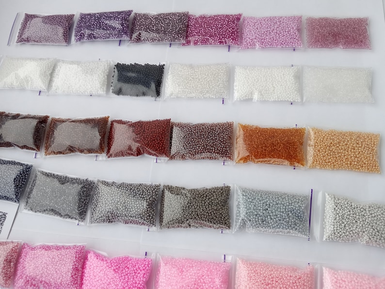 May include: Assortment of small, colorful beads in clear plastic bags. The beads are in various shades, including purple, white, black, brown, and gold. The bags are arranged in rows, creating a visually organized display of craft supplies.