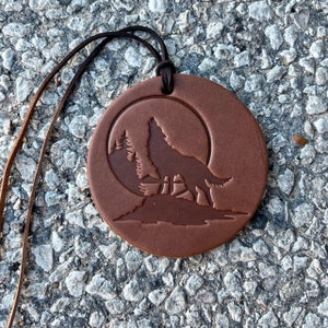 May include: A brown leather pendant with a wolf howling at the moon silhouette. The pendant is attached to a brown leather cord.