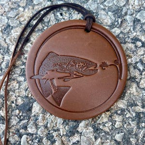May include: A circular brown leather pendant featuring an embossed design of a trout fish swimming toward a fishing lure. The pendant is suspended from a thin dark brown leather cord that passes through a small hole at the top of the disc.