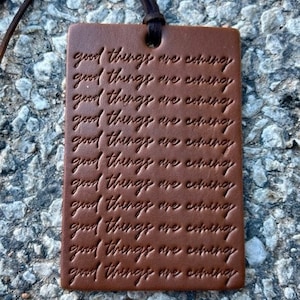 Puede incluir: Una etiqueta rectangular de cuero marrón con la frase en relieve "good things are coming" repetida varias veces. Una cuerda de cuero marrón oscuro está unida en la parte superior. La etiqueta es de un tono cálido y terroso.