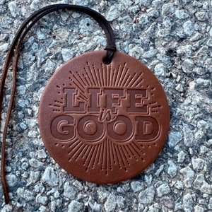May include: A round, brown leather ornament with the embossed text "LIFE is GOOD" in a stylized font. The design includes a sunburst pattern radiating from behind the text. A dark brown leather cord is attached for hanging.