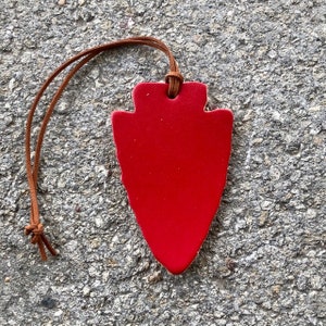 May include: A red arrowhead-shaped ornament with a brown leather cord. The ornament is made of ceramic and has a smooth, glossy finish.