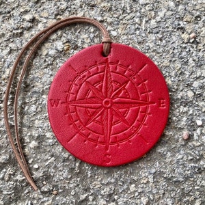 May include: A red leather compass charm with a brown leather cord. The compass features a detailed design with the cardinal directions marked.