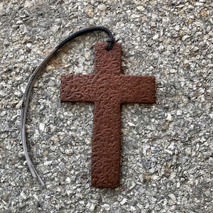 May include: A brown leather cross bookmark with a decorative floral pattern. The bookmark has a black leather strap for hanging.