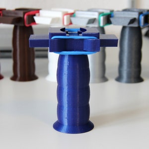 Könnte beinhalten: Ein blauer 3D-gedruckter Griff mit einem blauen Gummiband um die Oberseite. Der Griff ist von anderen Griffen in verschiedenen Farben umgeben, darunter rot, braun, weiß, grau und gelb.