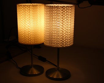 May include: Two table lamps with white patterned lampshades and silver bases. The lamps are turned on and casting a warm glow.
