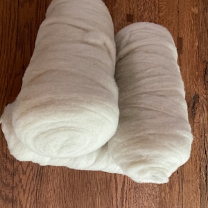 May include: Two rolls of white, unspun wool roving. The fibers are soft, fluffy, and densely packed into cylindrical shapes. The texture appears fibrous and airy, with a consistent off-white color throughout the material, showing natural variations.