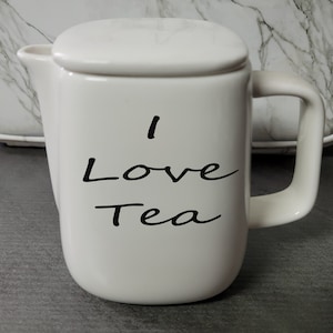 May include: White ceramic teapot with a lid and a handle. The teapot has black text that says "I Love Tea".