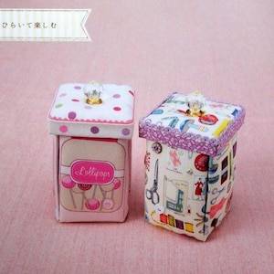 May include: Two decorative fabric boxes with lids. One box is pink with a lollipop design, the other is patterned with sewing-themed illustrations. Both have crystal-like knobs on top and are set against a pink background.
