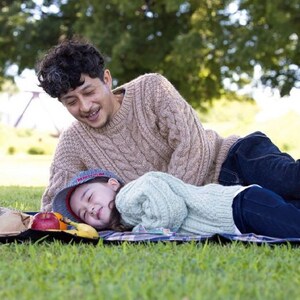 May include: A man and a young girl are lying on a blanket in a grassy field. The man is wearing a brown sweater and jeans. The girl is wearing a light green sweater and a blue and red hat. There are some fruit and bananas on the blanket.