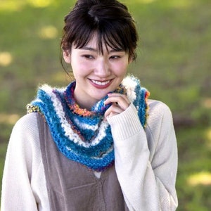 May include: A woman wearing a white sweater and a colorful crocheted scarf. The scarf has blue, white, and orange stripes.