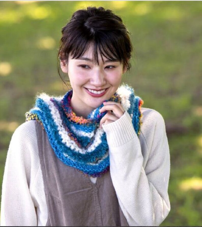 May include: A woman wearing a white sweater and a colorful crocheted scarf. The scarf has blue, white, and orange stripes.