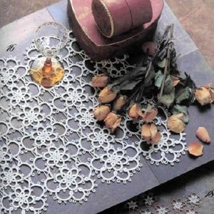 May include: A white crocheted doily with a delicate floral pattern. The doily is laid on a gray surface with dried roses and a small glass.