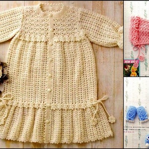 May include: A crocheted baby dress with long sleeves and a ruffled hem. The dress is cream colored and has a button closure down the front. The image also shows matching crocheted baby booties and a hat in pink and blue.