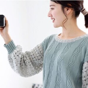 May include: A woman wearing a light blue and white knitted sweater with a wavy pattern. She is holding a black digital camera in her right hand and is looking at the camera.