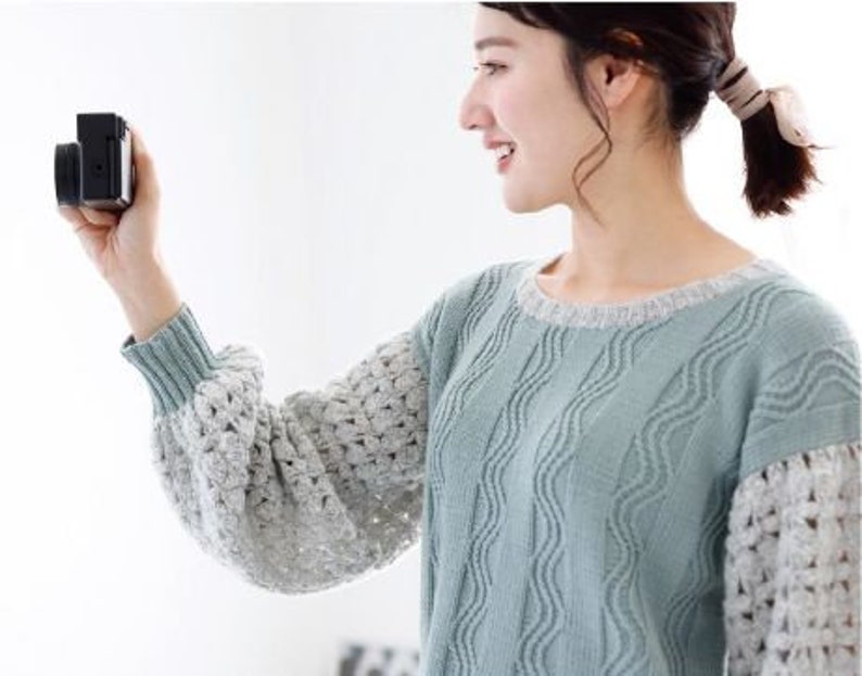 May include: A woman wearing a light blue and white knitted sweater with a wavy pattern. She is holding a black digital camera in her right hand and is looking at the camera.