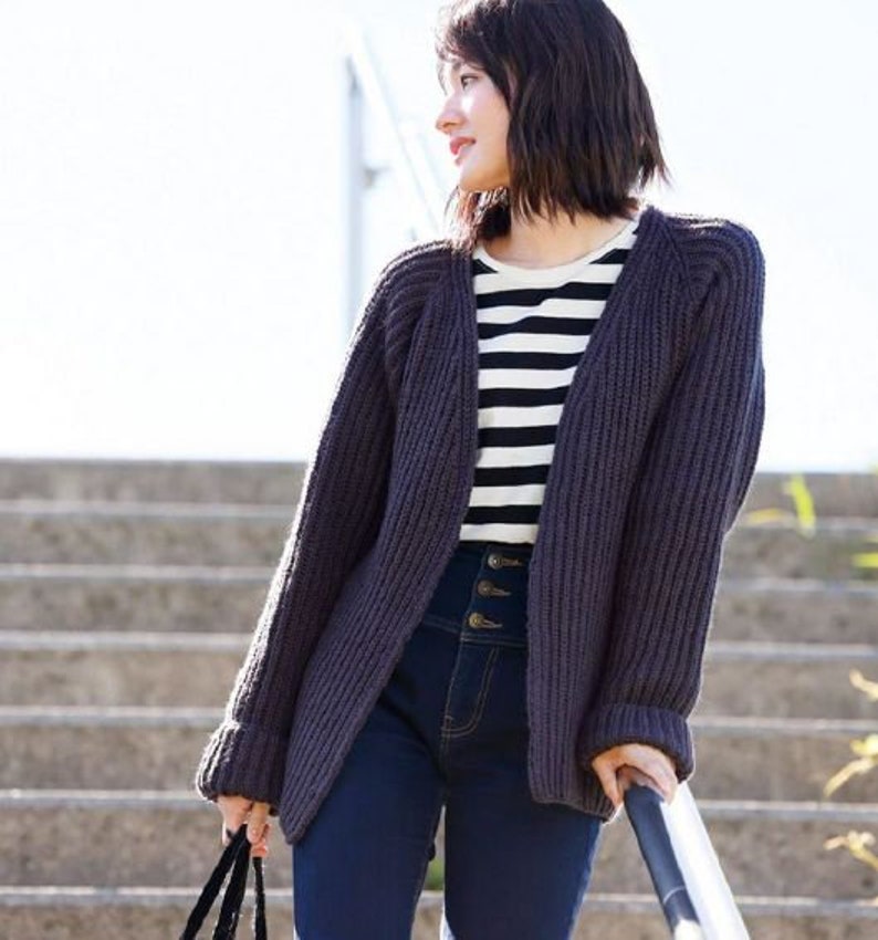 May include: A woman wearing a dark gray knitted cardigan with a striped shirt underneath and dark blue jeans. She is standing on a set of stairs and holding onto a railing.